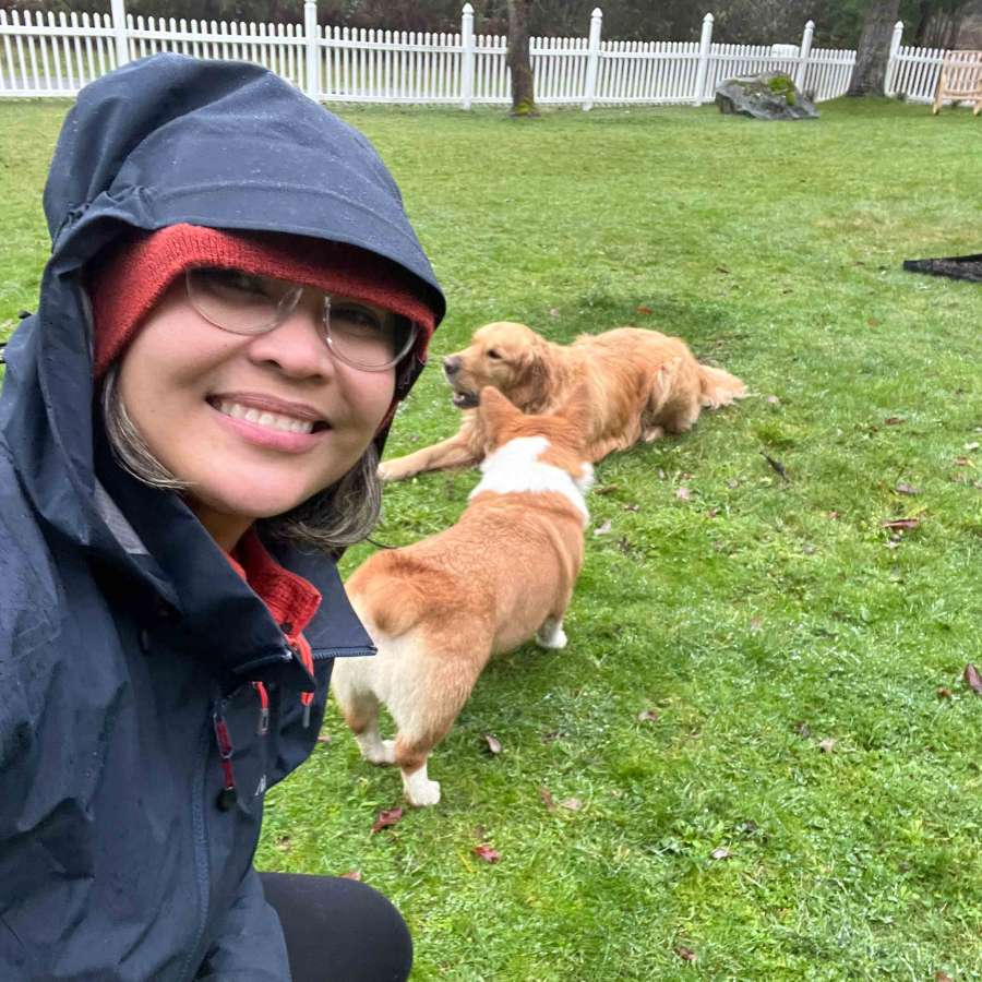 Luisa in rain jacket and glasses sitting on grass with two dogs